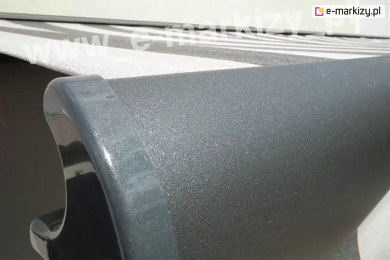 Zbliżenie belki przedniej markizy Palladio Aluprof wykonanej z ekstrudowanego aluminium malowanego proszkowo na kolor srebrzysty grafit oraz zaślepki bocznej z PCV w dopasowanym do aluminium kolorze. Nad belką widoczna tkanina w pasy o dwóch odcieniach szarości oraz jasna ściana, do której przymocowano markizę.