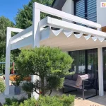Pergola drewniana kolumnowa Moza w białym kolorze konstrukcji z rozsuniętym systemem zadaszenia tkaninowego. Jasna tkanina dachu rzymskiego jest rozciągnięta pod konstrukcją, tworząc charakterystyczne, głębokie i miękko opadające fałdy w kształcie litery V, które są podwieszone na poziomych prowadnicach. Konstrukcja o białym wykończeniu kontrastuje z ciemnymi profilami okiennymi budynku oraz grafitowym zestawem mebli ogrodowych ustawionych pod zadaszeniem. Na tarasie z jasnych płyt gresowych widoczne jest nowoczesne wyposażenie, w tym sofa z poduszkami oraz rośliny doniczkowe w minimalistycznych, szarych pojemnikach. Na pierwszym planie dominuje dekoracyjnie przycięta sosna oraz krzewy na pniu, rosnące w pasie wysypanym białym grysem. Wzdłuż krawędzi tarasu rośnie lawenda oraz inne niskie krzewy ozdobne. Budynek posiada białą elewację z wstawką z desek elewacyjnych w kolorze ciepłego drewna, co koresponduje z naturalnymi elementami ogrodu. W tle, za konstrukcją pergoli, widoczna jest ściana wysokich, zielonych drzew liściastych oraz fragment bocznej ściany budynku z metalową rynną i osprzętem elektrycznym przy ziemi.
