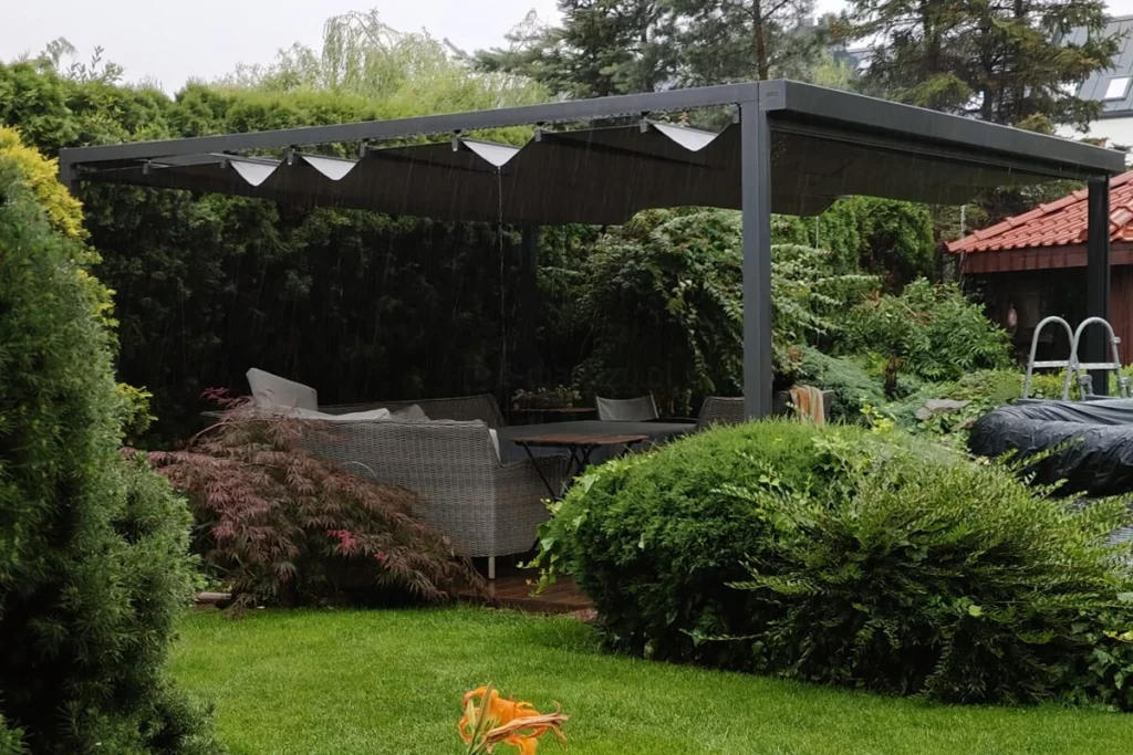 Nowoczesna pergola segmentowa Dragon w kolorze antracytowym zamontowana na drewnianym tarasie w otoczeniu gęstej roślinności ogrodowej podczas opadów deszczu. Konstrukcja pergoli opiera się na solidnych, kwadratowych profilach aluminiowych, które wspierają system ruchomego, rzymskiego zadaszenia. Tkanina dachowa w ciemnym odcieniu szarości jest częściowo rozsunięta, tworząc charakterystyczne, miękko układające się fałdy materiału podwieszone na poprzecznych belkach przeciwwietrznych. Zdjęcie prezentuje funkcjonalność odprowadzania wody, gdzie krople deszczu spływają z napiętego poszycia, chroniąc znajdujące się pod spodem meble ogrodowe z technorattanu oraz stolik kawowy. Całość konstrukcji jest wkomponowana w zielony krajobraz z wysokimi tujami, krzewami ozdobnymi oraz zadbanym trawnikiem, co podkreśla odporność materiałów na trudne warunki atmosferyczne. Widoczny jest boczny profil prowadnicy oraz system mocowania tkaniny, który zapewnia stabilność dachu nawet przy obciążeniu wilgocią. W tle widać fragmenty zabudowy sąsiedzkiej oraz czerwoną dachówkę altany, co osadza produkt w realnej aranżacji przydomowej strefy relaksu.
