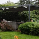 Nowoczesna pergola segmentowa Dragon w kolorze antracytowym zamontowana na drewnianym tarasie w otoczeniu gęstej roślinności ogrodowej podczas opadów deszczu. Konstrukcja pergoli opiera się na solidnych, kwadratowych profilach aluminiowych, które wspierają system ruchomego, rzymskiego zadaszenia. Tkanina dachowa w ciemnym odcieniu szarości jest częściowo rozsunięta, tworząc charakterystyczne, miękko układające się fałdy materiału podwieszone na poprzecznych belkach przeciwwietrznych. Zdjęcie prezentuje funkcjonalność odprowadzania wody, gdzie krople deszczu spływają z napiętego poszycia, chroniąc znajdujące się pod spodem meble ogrodowe z technorattanu oraz stolik kawowy. Całość konstrukcji jest wkomponowana w zielony krajobraz z wysokimi tujami, krzewami ozdobnymi oraz zadbanym trawnikiem, co podkreśla odporność materiałów na trudne warunki atmosferyczne. Widoczny jest boczny profil prowadnicy oraz system mocowania tkaniny, który zapewnia stabilność dachu nawet przy obciążeniu wilgocią. W tle widać fragmenty zabudowy sąsiedzkiej oraz czerwoną dachówkę altany, co osadza produkt w realnej aranżacji przydomowej strefy relaksu.