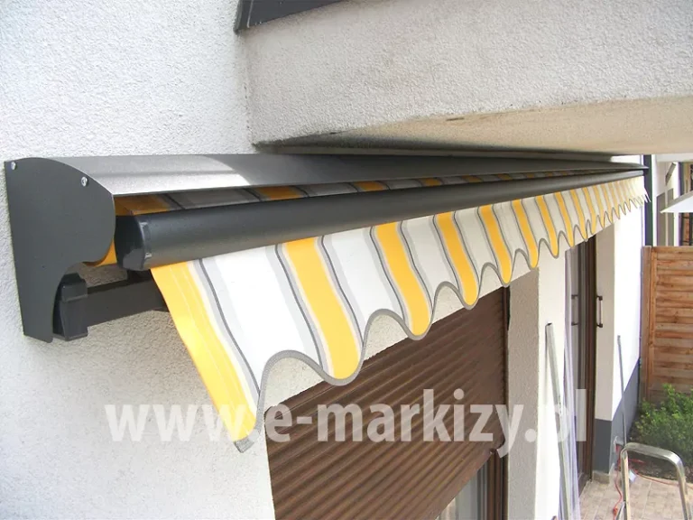 Markiza bez kasety Aluprof wyposażona w opcjonalny daszek do markiz 22,5cm w kolorze grafitowym, zawieszona pod balkonem na ścianie nad dwoma oknami tarasowymi. Widoczna falbana o falowanym kształcie w żółte, białe i szare pasy powiewa nad oknem osłoniętym roletą zewnętrzną w kolorze brązowym.