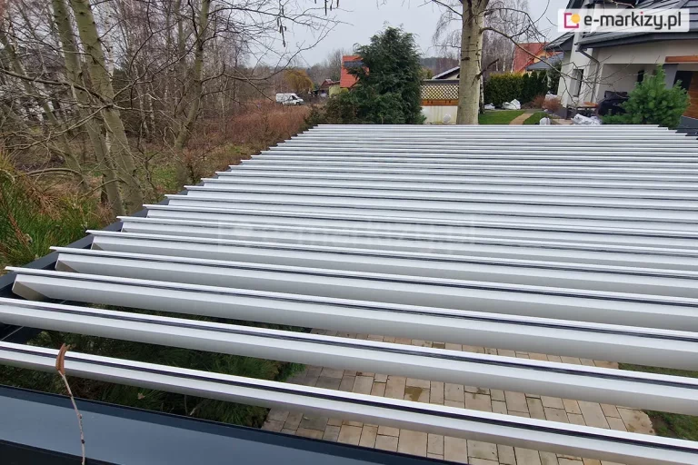 Zbliżenie na dach pergoli tarasowej z lotu ptaka, prezentujące system ruchomych lameli aluminiowych. Lamele w kolorze jasnoszarym są ustawione w pozycji lekko uchylonej, ukazując swoją budowę oraz czarne uszczelki na krawędziach. Rama zewnętrzna pergoli jest w kolorze antracytowym. Pod konstrukcją widoczny jest taras wyłożony prostokątną, jasną kostką. W dalszym planie rozpościera się widok na ogród z drzewami liściastymi bez liści, krzewy iglaste oraz sąsiednie zabudowania jednorodzinne z czerwonymi i szarymi dachami pod zachmurzonym niebem.