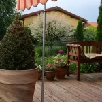 Na pierwszym planie ogromna donica z rośliną uformowaną w kształt stożka. Obok, po prawej widoczna podpora teleskopowa do markiz MOL, podtrzymująca pomarańczowo-czerwoną, falującą markizę nad drewnianym tarasem z donicami z roślinami oraz drewnianą ławką, w tle ogród i dom.