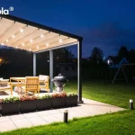 Widok z boku na podświetloną pergolę w aranżacji nocnej, zamontowaną na tarasie wyłożonym jasnymi płytami. Pergola tarasowa przyścienna tarasola Elegancy posiada Antracytową konstrukcję z rozłożonym, jasnym dachem tkaninowym jest intensywnie oświetlona przez zintegrowane punkty LED, które rzucają wyraźny blask na drewniane meble ogrodowe oraz czarny grill kulisty. Brzeg tarasu wyznacza szereg ciemnych donic z gęsto posadzonymi biało-różowymi kwiatami. Wzdłuż ścieżki prowadzącej do trawnika rozmieszczono niskie, nowoczesne słupki oświetleniowe. W tle widoczny jest ciemny ogród z zarysem domku dla dzieci oraz majaczące w oddali światła sąsiednich zabudowań pod nocnym, granatowym niebem.