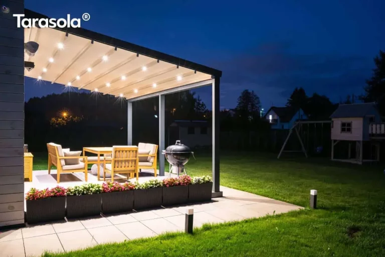Widok z boku na podświetloną pergolę w aranżacji nocnej, zamontowaną na tarasie wyłożonym jasnymi płytami. Pergola tarasowa przyścienna tarasola Elegancy posiada Antracytową konstrukcję z rozłożonym, jasnym dachem tkaninowym jest intensywnie oświetlona przez zintegrowane punkty LED, które rzucają wyraźny blask na drewniane meble ogrodowe oraz czarny grill kulisty. Brzeg tarasu wyznacza szereg ciemnych donic z gęsto posadzonymi biało-różowymi kwiatami. Wzdłuż ścieżki prowadzącej do trawnika rozmieszczono niskie, nowoczesne słupki oświetleniowe. W tle widoczny jest ciemny ogród z zarysem domku dla dzieci oraz majaczące w oddali światła sąsiednich zabudowań pod nocnym, granatowym niebem.