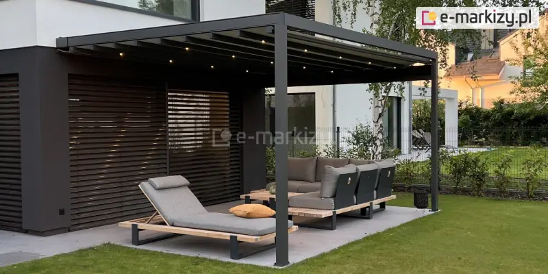 Nowoczesna przestrzeń wypoczynkowa pod pergolą, przy nowoczesnym domu o jasnej elewacji. Pergola tarasowa przyścienna Square Flat to prosty stelaż z grafitowego metalu z płaskim, belkowanym dachem. W górnej części konstrukcji zamontowane są niewielkie lampki LED, których ciepłe światło jest włączone. Pod pergolą ustawiono zestaw mebli ogrodowych w jednolitym, minimalistycznym stylu. Na pierwszym planie widoczny jest pojedynczy leżak z regulowanym zagłówkiem i jasnoszarą tapicerką, na którym leży ozdobna poduszka w kolorze musztardowym. Za nim znajduje się narożna sofa z miękkimi poduszkami i niskimi oparciami. Wszystkie meble mają drewniane ramy i czarne, metalowe nogi. Powierzchnia pod meblami wyłożona jest dużymi płytami tarasowymi, a cały taras przechodzi płynnie w zadbany, zielony trawnik. W tle widoczne są poziome żaluzje zewnętrzne na oknie, ozdobna zieleń ogrodu oraz fragment sąsiedniego budynku. Atmosfera miejsca jest nowoczesna i przytulna.
