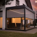 Nowoczesna pergola tarasowa Sb400 Aluprof w kolorze antracytowym, zamontowana przy białej elewacji piętrowego domu, sfotografowana o zmierzchu. Konstrukcja jest w pełni oświetlona zintegrowanymi listwami LED zamontowanymi wzdłuż wewnętrznej krawędzi dachu, co nadaje wnętrzu ciepły blask. Trzy boki pergoli są wyposażone w półtransparentne rolety zewnętrzne typu screen w kolorze beżowo-szarym, które są częściowo opuszczone. Pod zadaszeniem znajduje się zestaw wypoczynkowych mebli ogrodowych z technorattanu. Po lewej stronie widać wysokie, wąskie okno z opuszczoną roletą screen. Na zewnątrz, po prawej stronie tarasu, stoją dwie donice z podświetlanymi trawami ozdobnymi oraz wysoki świerk. Tło stanowi nastrojowe, ciemnopomarańczowe niebo po zachodzie słońca.