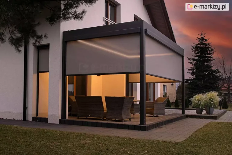 Nowoczesna pergola tarasowa Sb400 Aluprof w kolorze antracytowym, zamontowana przy białej elewacji piętrowego domu, sfotografowana o zmierzchu. Konstrukcja jest w pełni oświetlona zintegrowanymi listwami LED zamontowanymi wzdłuż wewnętrznej krawędzi dachu, co nadaje wnętrzu ciepły blask. Trzy boki pergoli są wyposażone w półtransparentne rolety zewnętrzne typu screen w kolorze beżowo-szarym, które są częściowo opuszczone. Pod zadaszeniem znajduje się zestaw wypoczynkowych mebli ogrodowych z technorattanu. Po lewej stronie widać wysokie, wąskie okno z opuszczoną roletą screen. Na zewnątrz, po prawej stronie tarasu, stoją dwie donice z podświetlanymi trawami ozdobnymi oraz wysoki świerk. Tło stanowi nastrojowe, ciemnopomarańczowe niebo po zachodzie słońca.