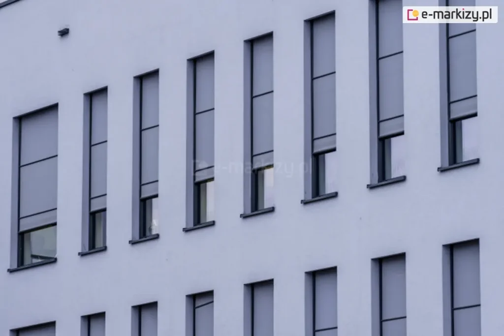 Elewacja nowoczesnego budynku z wąskimi, pionowymi oknami, na których zamontowano zewnętrzne rolety screenowe. Rolety Aluprof wykonane są z półprzezroczystej tkaniny w kolorze szarym i częściowo zasłaniają przeszklenia, pozostawiając dolne części okien odkryte. System prowadnic bocznych zapewnia stabilne prowadzenie tkaniny, co sugeruje zastosowanie rozwiązania typu ZIP. Kasety rolet są wbudowane i licują z elewacją, co zapewnia estetyczny i minimalistyczny wygląd fasady.