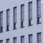 Elewacja nowoczesnego budynku z wąskimi, pionowymi oknami, na których zamontowano zewnętrzne rolety screenowe. Rolety Aluprof wykonane są z półprzezroczystej tkaniny w kolorze szarym i częściowo zasłaniają przeszklenia, pozostawiając dolne części okien odkryte. System prowadnic bocznych zapewnia stabilne prowadzenie tkaniny, co sugeruje zastosowanie rozwiązania typu ZIP. Kasety rolet są wbudowane i licują z elewacją, co zapewnia estetyczny i minimalistyczny wygląd fasady.