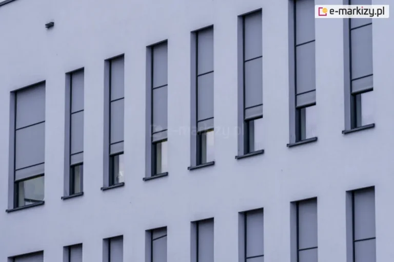Elewacja nowoczesnego budynku z wąskimi, pionowymi oknami, na których zamontowano zewnętrzne rolety screenowe. Rolety Aluprof wykonane są z półprzezroczystej tkaniny w kolorze szarym i częściowo zasłaniają przeszklenia, pozostawiając dolne części okien odkryte. System prowadnic bocznych zapewnia stabilne prowadzenie tkaniny, co sugeruje zastosowanie rozwiązania typu ZIP. Kasety rolet są wbudowane i licują z elewacją, co zapewnia estetyczny i minimalistyczny wygląd fasady.
