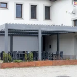 Duża, modułowa pergola tarasowa Sb400 w kolorze antracytowym, zainstalowana przy jasnej elewacji budynku jako zadaszenie ogródka restauracyjnego. Konstrukcja składa się z trzech segmentów, z których dwa są wyposażone w przesłony boczne (screeny) w kolorze szarym, zapewniające ochronę przed wiatrem i słońcem. Pod pergolą ustawiono liczne zestawy mebli gastronomicznych – czarne stoły i dopasowane do nich krzesła z tworzywa. Całość odgrodzona jest od placu z kostki brukowej długą, niską donicą wykonaną z drewna o ciepłym odcieniu, w której posadzono małe iglaki i ozdobne trawy. Na ścianie budynku powyżej pergoli widoczne są klasyczne okna w brązowych ramach oraz antena satelitarna. Zdjęcie prezentuje profesjonalną zabudowę przestrzeni komercyjnej, łączącą funkcję ochrony przed pogodą z estetycznym wydzieleniem strefy dla gości.