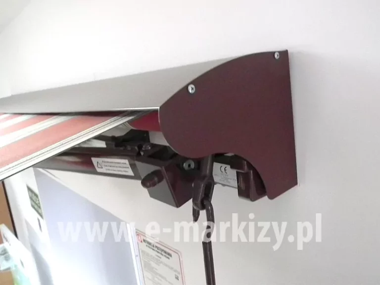 Zawieszona na białej ścianie nad otworem drzwiowym markiza z daszkiem uniwersalnym i napędem ręcznym w kolorze brązowym z rozwiniętym poszyciem w czerwono- biało-zielone pasy