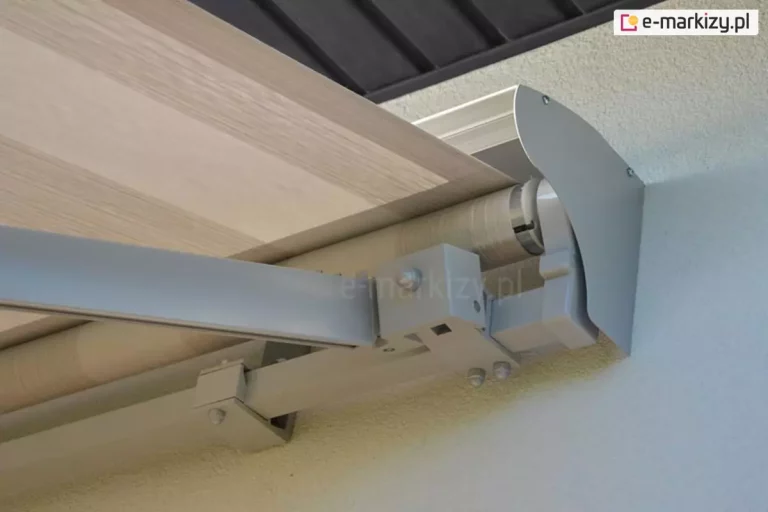 Techniczne zbliżenie na aluminiowy daszek ochronny, określany jako półkaseta, osłaniający mechanizm i tkaninę markizy tarasowej Jamaica firmy Aluprof. Konstrukcja w jasnoszarym, lakierowanym odcieniu jest zamontowana bezpośrednio do chropowatej, jasnej elewacji budynku pod podbitką dachową. Na zdjęciu widoczny jest boczny profil osłony o zaokrąglonym kształcie, przymocowany śrubami, który zabezpiecza rurę nawojową z nawiniętym materiałem. Poniżej daszka wysuwa się pasiaste poszycie markizy w tonacji beżu i szarości. W kadrze ukazano również detale konstrukcyjne systemu nośnego, w tym masywną kwadratową belkę montażową, solidne mocowania mechanizmu oraz fragment rozkładanego ramienia przegubowego, które odpowiada za stabilne naprężenie tkaniny. Fotografia przedstawia szczegóły techniczne produktu z perspektywy od dołu pod kątem.