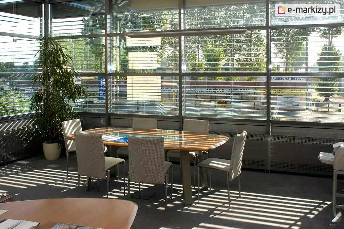 Wnętrze nowoczesnego biura lub sali konferencyjnej z dużymi przeszkleniami ściennymi, wyposażonymi w zewnętrzne żaluzje fasadowe wykonane z poziomych aluminiowych lameli, prawdopodobnie o profilu C80. Lamele ustawione są pod kątem umożliwiającym rozproszenie światła słonecznego, jednocześnie zapewniając widoczność na zewnątrz. Konstrukcja żaluzji oparta na prowadnicach bocznych, zamocowana na aluminiowej fasadzie przeszklonej. Na pierwszym planie znajduje się stół konferencyjny z blatem ze szkła hartowanego oraz sześć tapicerowanych krzeseł na metalowych nogach. Po lewej stronie widoczna wysoka roślina doniczkowa, z tyłu przez żaluzje widać zewnętrzną infrastrukturę – parking i banery reklamowe. Promienie słońca przechodzące przez lamele tworzą wyraźne cienie na podłodze i meblach. System żaluzji pełni funkcję ochrony przeciwsłonecznej, poprawia komfort pracy, ogranicza nagrzewanie wnętrza oraz chroni przed nadmiernym oświetleniem ekranu monitora.