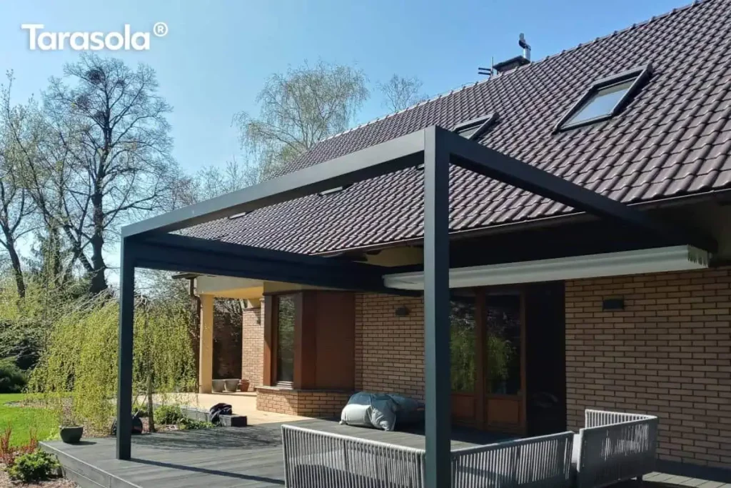 Zdjęcie prezentuje nowoczesną, prostokątną pergolę tarasową Tarasola Flat o konstrukcji aluminiowej w kolorze antracytowym, zamontowaną jako zadaszenie przestronnego tarasu wykonanego z ciemnych desek kompozytowych. Konstrukcja o minimalistycznym designie jest zintegrowana z elewacją parterowego budynku wykonanego z jasnobrązowej cegły klinkierowej i przykrytego spadzistym dachem z ciemnej dachówki. Pod pergolą znajduje się zestaw mebli wypoczynkowych z szarego technorattanu lub sznurka, składający się z narożnika z miękkimi poduszkami. Dach tkaninowy pergoli w kolorze białym jest obecnie całkowicie zwinięty w stronę elewacji, co odsłania widok na niebo i wpuszcza pełne światło na taras. W tle widoczny jest zadbany, zielony ogród z dużymi drzewami liściastymi, w tym wierzbą płaczącą, oraz fragment trawnika pod bezchmurnym niebem. Całość kompozycji podkreśla nowoczesną architekturę domu i funkcjonalne zagospodarowanie strefy relaksu na świeżym powietrzu.