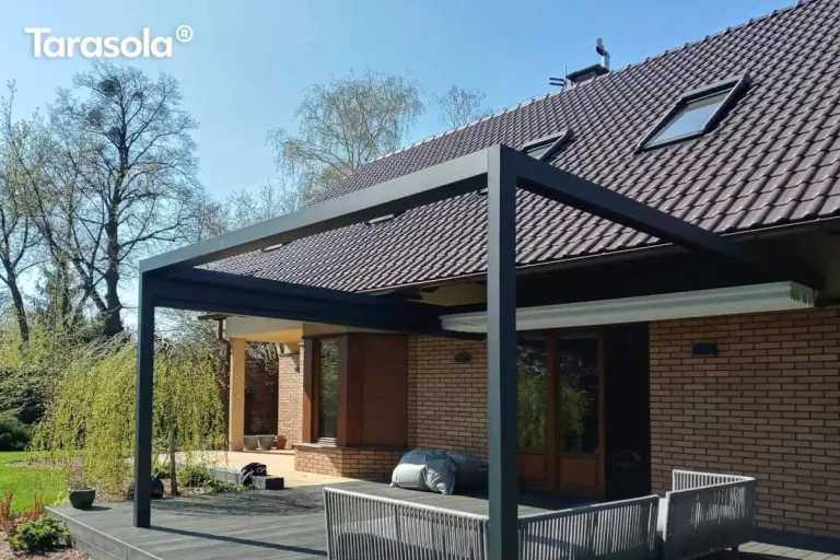 Zdjęcie prezentuje nowoczesną, prostokątną pergolę tarasową Tarasola Flat o konstrukcji aluminiowej w kolorze antracytowym, zamontowaną jako zadaszenie przestronnego tarasu wykonanego z ciemnych desek kompozytowych. Konstrukcja o minimalistycznym designie jest zintegrowana z elewacją parterowego budynku wykonanego z jasnobrązowej cegły klinkierowej i przykrytego spadzistym dachem z ciemnej dachówki. Pod pergolą znajduje się zestaw mebli wypoczynkowych z szarego technorattanu lub sznurka, składający się z narożnika z miękkimi poduszkami. Dach tkaninowy pergoli w kolorze białym jest obecnie całkowicie zwinięty w stronę elewacji, co odsłania widok na niebo i wpuszcza pełne światło na taras. W tle widoczny jest zadbany, zielony ogród z dużymi drzewami liściastymi, w tym wierzbą płaczącą, oraz fragment trawnika pod bezchmurnym niebem. Całość kompozycji podkreśla nowoczesną architekturę domu i funkcjonalne zagospodarowanie strefy relaksu na świeżym powietrzu.