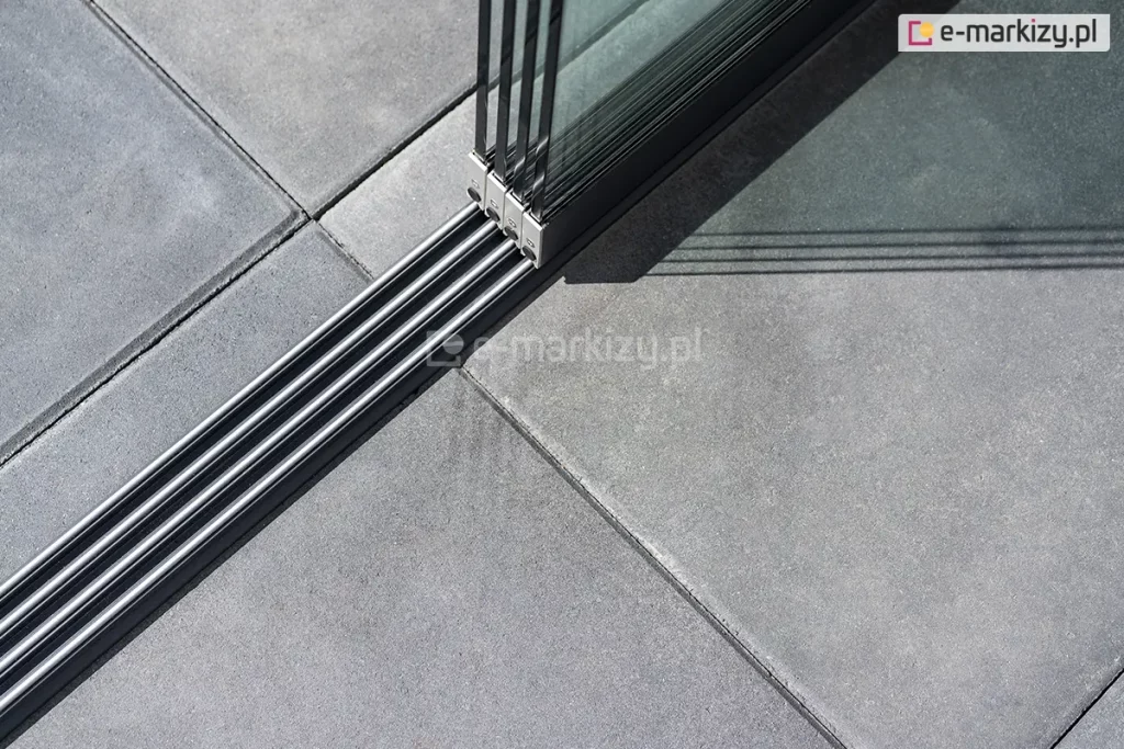 Zamocowane do kwadratowych betonowych płyt prowadnice 4-torowe do paneli Auris w kolorze antracytowym oraz osadzone na nich cztery panele szklane przesuwne tworzące zabudowę tarasu.