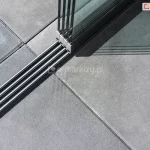 Zamocowane do kwadratowych betonowych płyt prowadnice 4-torowe do paneli Auris w kolorze antracytowym oraz osadzone na nich cztery panele szklane przesuwne tworzące zabudowę tarasu.