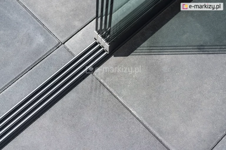 Zamocowane do kwadratowych betonowych płyt prowadnice 4-torowe do paneli Auris w kolorze antracytowym oraz osadzone na nich cztery panele szklane przesuwne tworzące zabudowę tarasu.