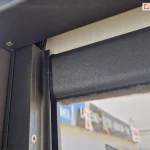 Słupek konstrukcji pergoli sb 400 Aluprof w kolorze antracytowym z zamontowaną refleksolą ziiip 120 umieszczoną w prowadnicy bocznej. Konstrukcja refleksola dopasowana została do koloru pergoli, w belce dolnej widoczna uszczelka szczotkowa oraz zwinięta tkanina screen w kolorze beżowym.