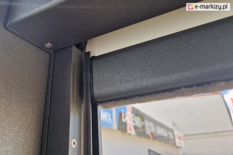 Słupek konstrukcji pergoli sb 400 Aluprof w kolorze antracytowym z zamontowaną refleksolą ziiip 120 umieszczoną w prowadnicy bocznej. Konstrukcja refleksola dopasowana została do koloru pergoli, w belce dolnej widoczna uszczelka szczotkowa oraz zwinięta tkanina screen w kolorze beżowym.