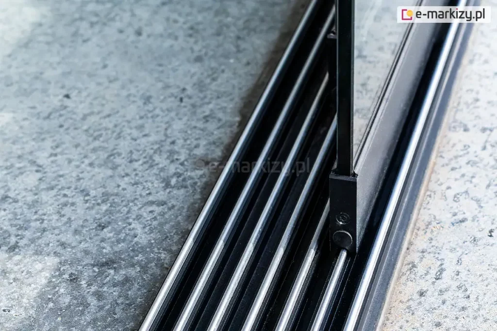 Zbliżenie na dolną, aluminiową prowadnicę wielotorową przeznaczoną do szklanych drzwi przesuwnych Auris, zamontowaną na betonowym podłożu. Antracytowe prowadnice 7-torowe do paneli Auris wyposażone są w system równoległych, srebrnych szyn o półokrągłym przekroju, po których poruszają się wózki jezdne. Na jednej z wewnętrznych szyn widoczny jest fragment dolnego profilu przesuwnego skrzydła drzwiowego w kolorze czarnym, w którym osadzona jest pionowa szyba. Konstrukcja profilu skrzydła idealnie dopasowuje się do wypukłego kształtu szyny, zapewniając stabilne prowadzenie elementu szklanego. Po lewej i prawej stronie prowadnicy znajduje się surowa, szara nawierzchnia betonowa o nieregularnej strukturze.