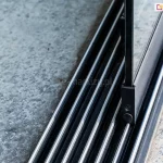 Zbliżenie na dolną, aluminiową prowadnicę wielotorową przeznaczoną do szklanych drzwi przesuwnych Auris, zamontowaną na betonowym podłożu. Antracytowe prowadnice 7-torowe do paneli Auris wyposażone są w system równoległych, srebrnych szyn o półokrągłym przekroju, po których poruszają się wózki jezdne. Na jednej z wewnętrznych szyn widoczny jest fragment dolnego profilu przesuwnego skrzydła drzwiowego w kolorze czarnym, w którym osadzona jest pionowa szyba. Konstrukcja profilu skrzydła idealnie dopasowuje się do wypukłego kształtu szyny, zapewniając stabilne prowadzenie elementu szklanego. Po lewej i prawej stronie prowadnicy znajduje się surowa, szara nawierzchnia betonowa o nieregularnej strukturze.