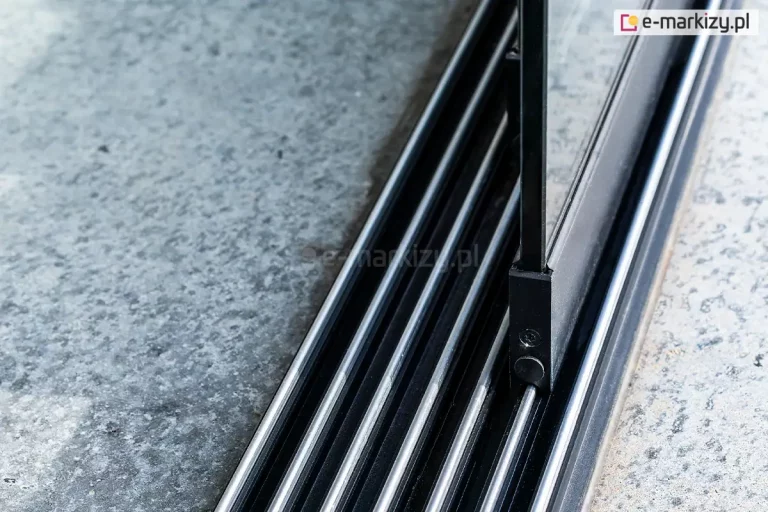 Zbliżenie na dolną, aluminiową prowadnicę wielotorową przeznaczoną do szklanych drzwi przesuwnych Auris, zamontowaną na betonowym podłożu. Antracytowe prowadnice 7-torowe do paneli Auris wyposażone są w system równoległych, srebrnych szyn o półokrągłym przekroju, po których poruszają się wózki jezdne. Na jednej z wewnętrznych szyn widoczny jest fragment dolnego profilu przesuwnego skrzydła drzwiowego w kolorze czarnym, w którym osadzona jest pionowa szyba. Konstrukcja profilu skrzydła idealnie dopasowuje się do wypukłego kształtu szyny, zapewniając stabilne prowadzenie elementu szklanego. Po lewej i prawej stronie prowadnicy znajduje się surowa, szara nawierzchnia betonowa o nieregularnej strukturze.