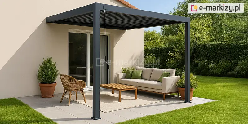 Nowoczesna pergola lamelowa przyścienna Marlo w kolorze antracytowym, zamontowana do elewacji budynku o jasnym, beżowym wykończeniu. Konstrukcja zadaszenia opiera się na dwóch solidnych profilach przednich, a dach składa się z poziomych lameli ustawionych w pozycji zamkniętej. Pod pergolą zaaranżowano strefę wypoczynkową na tarasie wyłożonym jasnymi płytami, na którą składają się: jasnobeżowa sofa z zielonymi poduszkami, drewniany stolik kawowy, pleciony fotel oraz dywan zewnętrzny. Po bokach ustawiono rośliny w glinianych donicach, a całość otacza zadbany trawnik oraz gęsty, zielony żywopłot. Przy przednim słupie widoczna jest ręczna korba służąca do regulacji kąta nachylenia lameli.
