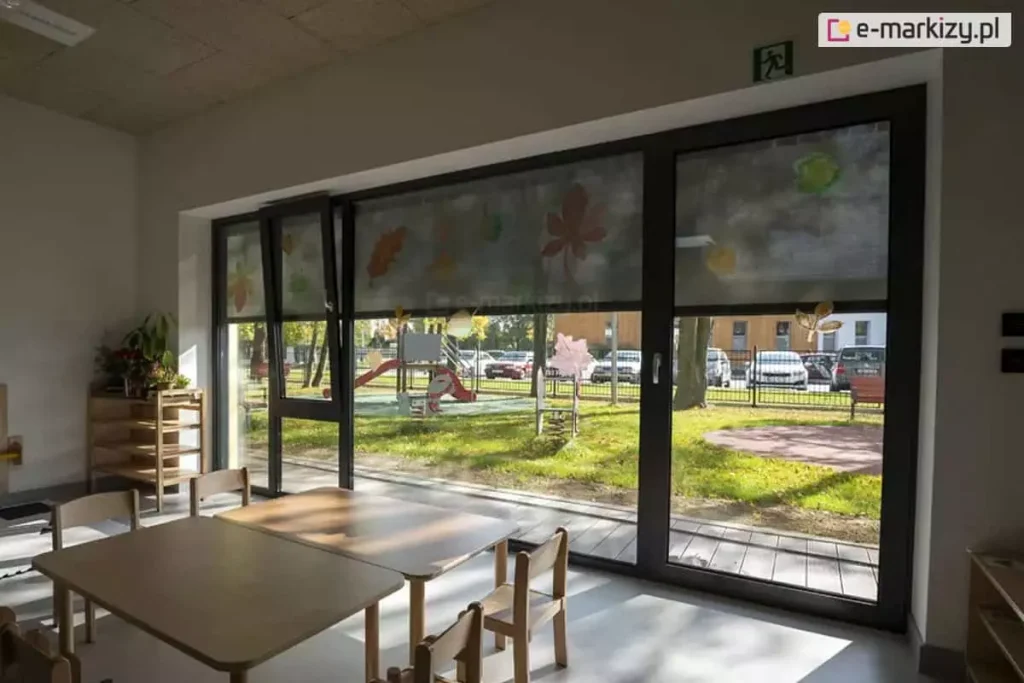 Wnętrze nowocześnie urządzonej sali przedszkolnej wypełnia naturalne światło przenikające przez duże, przeszklone okna w grafitowych ramach, z których jedno po lewej stronie jest uchylone. Górną część przeszkleń częściowo zasłaniają opuszczone, półprzezroczyste rolety screen, na których od wewnętrznej strony umieszczono ozdobne wycinanki z papieru przedstawiające kolorowe liście i motywy jesienne. Przez zasłonięte i odsłonięte fragmenty okien doskonale widać zewnętrzny plac zabaw z kolorowymi zjeżdżalniami, bujakami na sprężynach, zieloną trawą, chodnikiem oraz zaparkowanymi w tle samochodami osobowymi. Na jasnej podłodze w pomieszczeniu ustawiono niskie, drewniane stoliki o zaokrąglonych rogach przeznaczone dla dzieci, wokół których rozstawiono dopasowane rozmiarem małe krzesełka. Przy lewej ścianie znajduje się niski regał z zabawkami oraz ozdobne rośliny doniczkowe ustawione na blacie, a nad oknem zamontowano zielony znak ewakuacyjny wskazujący kierunek wyjścia. Całość tworzy przyjazną, funkcjonalną i dobrze doświetloną przestrzeń edukacyjną, w której nowoczesne osłony okienne skutecznie regulują dopływ światła słonecznego, zapewniając komfort przebywającym w środku maluchom.