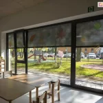 Wnętrze nowocześnie urządzonej sali przedszkolnej wypełnia naturalne światło przenikające przez duże, przeszklone okna w grafitowych ramach, z których jedno po lewej stronie jest uchylone. Górną część przeszkleń częściowo zasłaniają opuszczone, półprzezroczyste rolety screen, na których od wewnętrznej strony umieszczono ozdobne wycinanki z papieru przedstawiające kolorowe liście i motywy jesienne. Przez zasłonięte i odsłonięte fragmenty okien doskonale widać zewnętrzny plac zabaw z kolorowymi zjeżdżalniami, bujakami na sprężynach, zieloną trawą, chodnikiem oraz zaparkowanymi w tle samochodami osobowymi. Na jasnej podłodze w pomieszczeniu ustawiono niskie, drewniane stoliki o zaokrąglonych rogach przeznaczone dla dzieci, wokół których rozstawiono dopasowane rozmiarem małe krzesełka. Przy lewej ścianie znajduje się niski regał z zabawkami oraz ozdobne rośliny doniczkowe ustawione na blacie, a nad oknem zamontowano zielony znak ewakuacyjny wskazujący kierunek wyjścia. Całość tworzy przyjazną, funkcjonalną i dobrze doświetloną przestrzeń edukacyjną, w której nowoczesne osłony okienne skutecznie regulują dopływ światła słonecznego, zapewniając komfort przebywającym w środku maluchom.