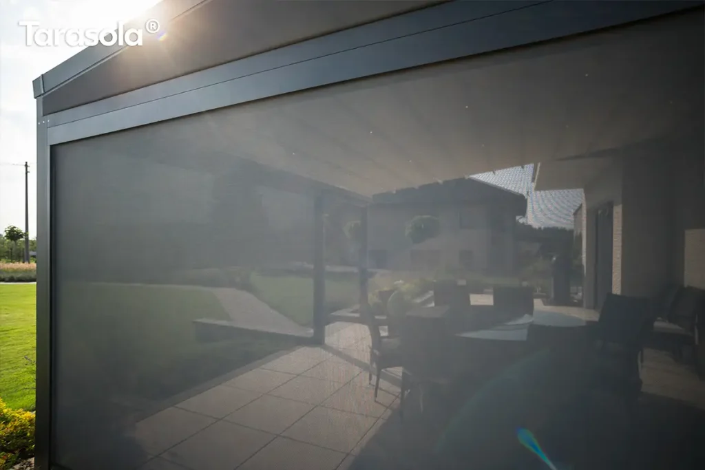 Szczegółowe ujęcie zadaszenia tarasowego typu pergola z zamontowanymi osłonami bocznymi w postaci rolet typu screen, przedstawione od wewnątrz z widokiem na ogród. W górnej części widoczny jest sufit z paneli aluminiowych z wbudowanym oświetleniem punktowym LED rozmieszczonym symetrycznie w kilku rzędach, emitującym ciepłe, nastrojowe światło. Po bokach i z przodu konstrukcji opuszczone są przezierne rolety materiałowe w ciemnym kolorze, które skutecznie zacieniają wnętrze, jednocześnie zachowując widoczność zewnętrzną. Przez strukturalną tkaninę rolet przebijają się promienie zachodzącego słońca, tworząc efektowne refleksy świetlne. W dolnej części kadru znajduje się nowoczesna balustrada lub murek tarasowy wykończony ciemnymi płytami, a w tle rozciąga się zielony ogród z równo przyciętymi iglakami i widocznym ogrodzeniem. W lewym górnym rogu widoczne jest białe logo producenta.