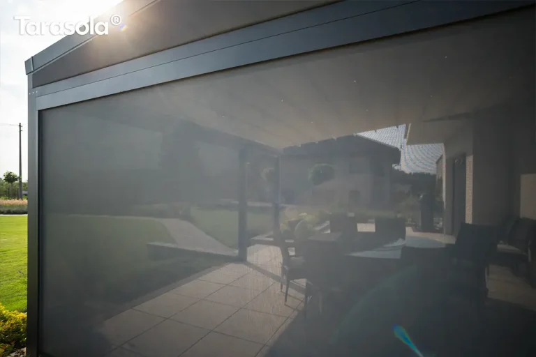 Szczegółowe ujęcie zadaszenia tarasowego typu pergola z zamontowanymi osłonami bocznymi w postaci rolet typu screen, przedstawione od wewnątrz z widokiem na ogród. W górnej części widoczny jest sufit z paneli aluminiowych z wbudowanym oświetleniem punktowym LED rozmieszczonym symetrycznie w kilku rzędach, emitującym ciepłe, nastrojowe światło. Po bokach i z przodu konstrukcji opuszczone są przezierne rolety materiałowe w ciemnym kolorze, które skutecznie zacieniają wnętrze, jednocześnie zachowując widoczność zewnętrzną. Przez strukturalną tkaninę rolet przebijają się promienie zachodzącego słońca, tworząc efektowne refleksy świetlne. W dolnej części kadru znajduje się nowoczesna balustrada lub murek tarasowy wykończony ciemnymi płytami, a w tle rozciąga się zielony ogród z równo przyciętymi iglakami i widocznym ogrodzeniem. W lewym górnym rogu widoczne jest białe logo producenta.