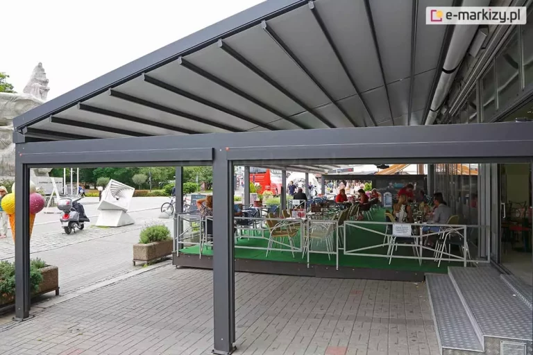 Zdjęcie prezentuje perspektywiczne ujęcie zewnętrznego ogródka restauracyjnego zadaszonego wielosegmentową pergolą tarasową model Solid Selt. Konstrukcja aluminiowa w kolorze ciemnego antracytu składa się z szeregu masywnych słupów frontowych połączonych poziomymi belkami, tworząc stabilny szkielet dla ruchomego dachu. Zadaszenie wykonane z jasnej, technicznej tkaniny jest w pełni rozłożone i wspiera się na poprzecznych profilach aluminiowych rozmieszczonych w regularnych odstępach. Pod pergolą, na podłożu wyłożonym zieloną wykładziną, ustawione są liczne stoliki restauracyjne z jasnymi krzesłami, przy których siedzą goście. Ogródek jest oddzielony od publicznego chodnika niską, białą barierka o nowoczesnym, geometrycznym wzorze. Na pierwszym tle pomiędzy podporami pergoli widoczne są skrzynki zwiniętych refleksoli ziiip. W tle widoczna jest przeszklona fasada lokalu oraz fragmenty architektury miejskiej, w tym rzeźby i elementy zieleni publicznej. Zdjęcie zostało wykonane w świetle dziennym, co uwydatnia dużą powierzchnię osłony przeciwsłonecznej zapewnianej przez system Solid.