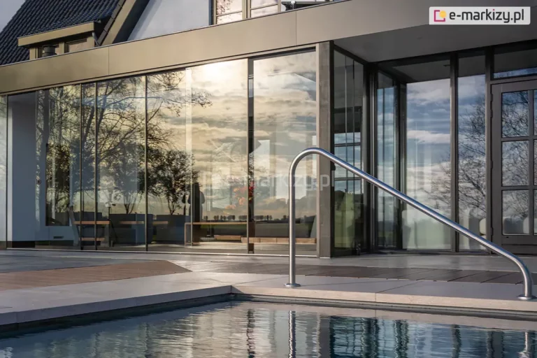 Fotografia prezentująca nowoczesną architekturę zewnętrzną z wykorzystaniem bezramowego systemu przesuwnych drzwi szklanych Auris, które zamykają przestronny salon ogrodowy zintegrowany z bryłą domu. Wielkoformatowe, szklane panele ze szkła bezpiecznego tworzą gładką, przezroczystą ścianę, w której efektownie odbija się popołudniowe słońce przedzierające się przez malownicze chmury oraz zarysy drzew w tle. Na pierwszym planie, przed przeszkloną fasadą, widoczny jest fragment luksusowego basenu z czystą, błękitną wodą oraz metalowa, wygięta poręcz ułatwiająca wchodzenie do wody. Otoczenie basenu wykończone jest jasnoszarymi płytami tarasowymi oraz pasem z ciemnego drewna lub kompozytu. Wewnątrz przeszklonej konstrukcji subtelnie rysują się zarysy luksusowych mebli wypoczynkowych. Zdjęcie wykonane pod niskim kątem w ciepłym świetle dnia podkreśla ekskluzywny design, elegancję i transparentność całego systemu przeszkleń.