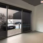 Wysokie okna z opuszczonymi roletami screen w przestrzeni biurowej, półprzezroczysta tkanina pokazuje zarysy aut i zimowego krajobrazu. W głębi po prawej stronie widoczne wejście do budynku z nagrzewnicą nad drzwiami oraz nieco bliżej ustawiony stolik z fotelem.