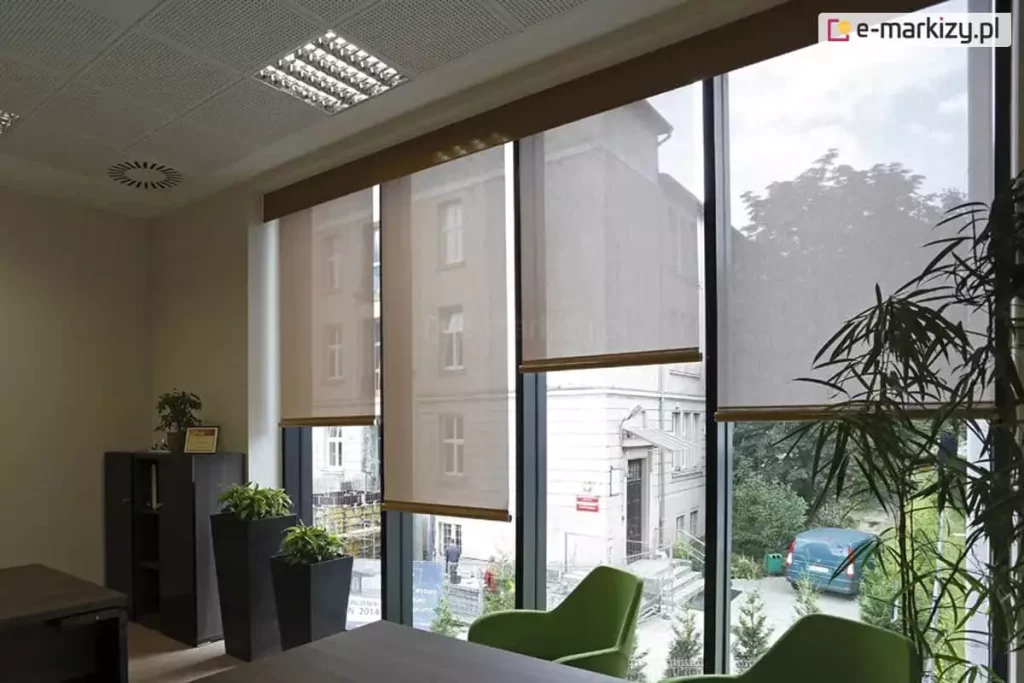 Widok wnętrza nowoczesnego biura na system czterech refleksoli XL, zamontowanych na wysokim przeszkleniu z ciemnymi profilami. Rolety o beżowej tkaninie technicznej są ustawione na różnych wysokościach, prezentując funkcjonalność osłon w zakresie selektywnego zacieniania pomieszczenia. Półtransparentny materiał o drobnym splocie pozwala na obserwację otoczenia zewnętrznego, w tym klasycznej kamienicy, parkingu oraz zieleni miejskiej, jednocześnie chroniąc wnętrze przed bezpośrednim promieniowaniem słonecznym. W pomieszczeniu widoczne są nowoczesne, zielone fotele biurowe, wysokie czarne donice z roślinami ozdobnymi oraz fragment biurka. Sufit kasetonowy wyposażony jest w rastrowe oprawy oświetleniowe oraz okrągły nawiewnik systemu wentylacji, a naturalne światło dzienne miękko rozprasza się na tkaninie rolet, tworząc komfortowe warunki do pracy biurowej.