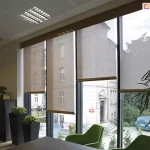 Widok wnętrza nowoczesnego biura na system czterech refleksoli XL, zamontowanych na wysokim przeszkleniu z ciemnymi profilami. Rolety o beżowej tkaninie technicznej są ustawione na różnych wysokościach, prezentując funkcjonalność osłon w zakresie selektywnego zacieniania pomieszczenia. Półtransparentny materiał o drobnym splocie pozwala na obserwację otoczenia zewnętrznego, w tym klasycznej kamienicy, parkingu oraz zieleni miejskiej, jednocześnie chroniąc wnętrze przed bezpośrednim promieniowaniem słonecznym. W pomieszczeniu widoczne są nowoczesne, zielone fotele biurowe, wysokie czarne donice z roślinami ozdobnymi oraz fragment biurka. Sufit kasetonowy wyposażony jest w rastrowe oprawy oświetleniowe oraz okrągły nawiewnik systemu wentylacji, a naturalne światło dzienne miękko rozprasza się na tkaninie rolet, tworząc komfortowe warunki do pracy biurowej.