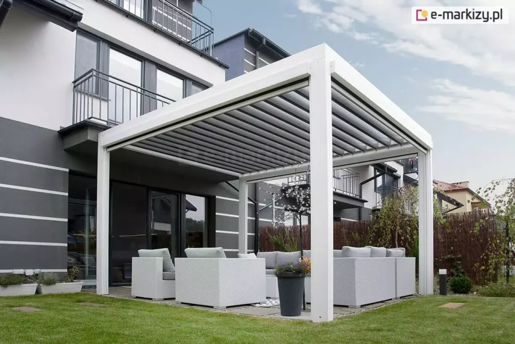 Pergola w kolorze białym - model SB400 Aluprof ustawiona w ogrodzie w sąsiedztwie domków o biało-szarej kolorystyce, na zielonym trawniku. Po pergolą ustawiono meble wypoczynkowe z rattanu malowanego na biało z poduchami w jasnoszarym odcieniu. Pod dachem znalazła się także lampka w kształcie drzewka z licznymi punktami świetlnymi na gałązkach oraz duża donica czarna z kwitnącymi roślinami.