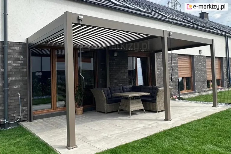 Duża pergola tarasowa przyścienna w kolorze beżowo-szarym (taupe), zamontowana przy elewacji nowoczesnego domu jednorodzinnego. Konstrukcja opiera się na trzech masywnych słupach frontowych i składa się z dwóch sekcji dachu lamelowego. Lewa sekcja dachu ma lamele ustawione pod kątem, co pozwala na dostęp światła i wentylację, natomiast prawa sekcja pozostaje zamknięta. Pod zadaszeniem, na tarasie wyłożonym wielkoformatowymi, jasnoszarymi płytami, ustawiono narożnik z technorattanu z ciemnymi poduszkami oraz dopasowany stolik. Na słupkach konstrukcyjnych widoczne są dwa oddzielne mechanizmy korbowe do niezależnej regulacji obu części dachu. Budynek posiada elewację z ciemnej cegły klinkierowej w dolnej części i jasnego tynku powyżej, a okna wyposażone są w zewnętrzne rolety w kolorze brązowym.