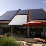 Szerokie ujęcie elewacji tylnej domu jednorodzinnego z dachem dwuspadowym pokrytym czerwoną dachówką ceramiczną, na którym zainstalowano rozbudowany system paneli fotowoltaicznych oraz nowoczesną roletę veranda V-Ziiip Aluprof. Veranda z tkaniną w kolorze jasnoszarym jest całkowicie rozwinięta i zamontowana bezpośrednio nad przeszklonym wykuszem lub ogrodem zimowym o ciemnych profilach, stanowiąc integralną część połaci dachowej. Po obu stronach markizy znajdują się liczne rzędy ciemnych modułów fotowoltaicznych, które wypełniają niemal całą dostępną powierzchnię dachu. Przed budynkiem rozciąga się przestronny taras wykonany z ciemnego drewna, na którym ustawiono meble ogrodowe, stół oraz leżaki z pomarańczowymi materacami. Czerwony parasol ogrodowy rozłożony nad stołem koresponduje kolorystycznie z barwą dachówki. Teren wokół tarasu jest bogato obsadzony zielenią, w tym wysokimi trawami ozdobnymi i krzewami, co nadaje posesji przytulny charakter. Całość sceny jest oświetlona jasnym, popołudniowym światłem przy bezchmurnym, błękitnym niebie, co podkreśla nowoczesną architekturę i proekologiczne rozwiązania zastosowane w budynku.