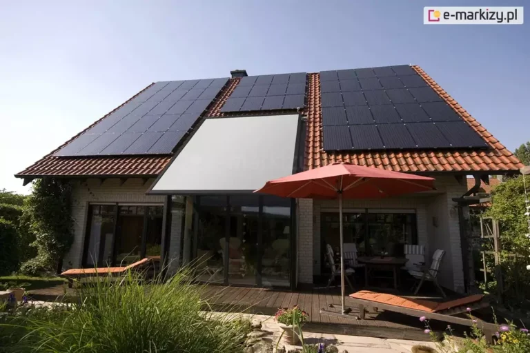 Szerokie ujęcie elewacji tylnej domu jednorodzinnego z dachem dwuspadowym pokrytym czerwoną dachówką ceramiczną, na którym zainstalowano rozbudowany system paneli fotowoltaicznych oraz nowoczesną roletę veranda V-Ziiip Aluprof. Veranda z tkaniną w kolorze jasnoszarym jest całkowicie rozwinięta i zamontowana bezpośrednio nad przeszklonym wykuszem lub ogrodem zimowym o ciemnych profilach, stanowiąc integralną część połaci dachowej. Po obu stronach markizy znajdują się liczne rzędy ciemnych modułów fotowoltaicznych, które wypełniają niemal całą dostępną powierzchnię dachu. Przed budynkiem rozciąga się przestronny taras wykonany z ciemnego drewna, na którym ustawiono meble ogrodowe, stół oraz leżaki z pomarańczowymi materacami. Czerwony parasol ogrodowy rozłożony nad stołem koresponduje kolorystycznie z barwą dachówki. Teren wokół tarasu jest bogato obsadzony zielenią, w tym wysokimi trawami ozdobnymi i krzewami, co nadaje posesji przytulny charakter. Całość sceny jest oświetlona jasnym, popołudniowym światłem przy bezchmurnym, błękitnym niebie, co podkreśla nowoczesną architekturę i proekologiczne rozwiązania zastosowane w budynku.