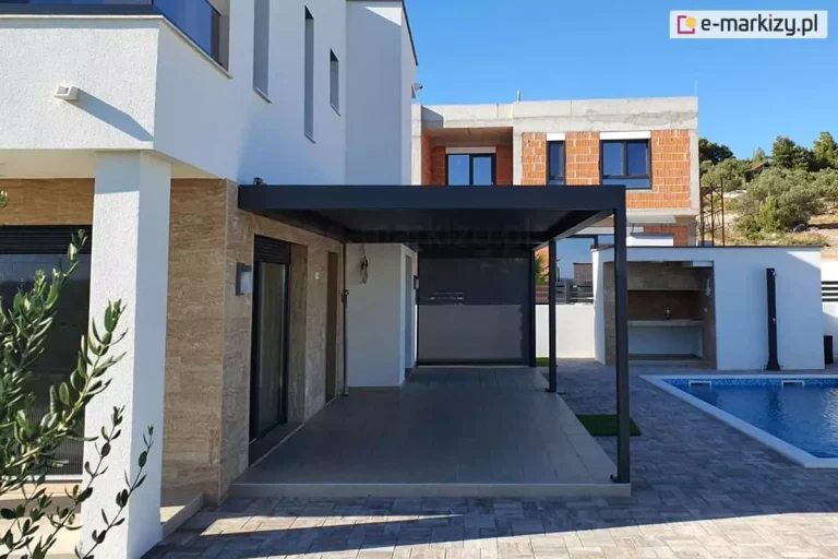 Nowoczesna pergola tarasowa Sb400 Aluprof o konstrukcji aluminiowej w kolorze antracytowym, zamontowana do elewacji luksusowej willi. Budynek posiada białą elewację połączoną z fragmentami wyłożonymi jasnobrązowym kamieniem dekoracyjnym. Pergola osłania przejście wzdłuż domu, a jej tylna część jest wyposażona w opuszczoną szarą roletę boczną typu screen. Podłoże tarasu wykonane jest z dużych, beżowych płyt, które płynnie przechodzą w plac wyłożony szarą kostką brukową. W tle po prawej stronie znajduje się niebieski basen ogrodowy oraz letnia kuchnia z wbudowanym grillem. W oddali widoczny jest sąsiedni budynek w stanie surowym z czerwonej cegły oraz zielone wzgórza pod bezchmurnym, błękitnym niebem. Na pierwszym planie po lewej stronie widać fragment gałęzi drzewa oliwnego.