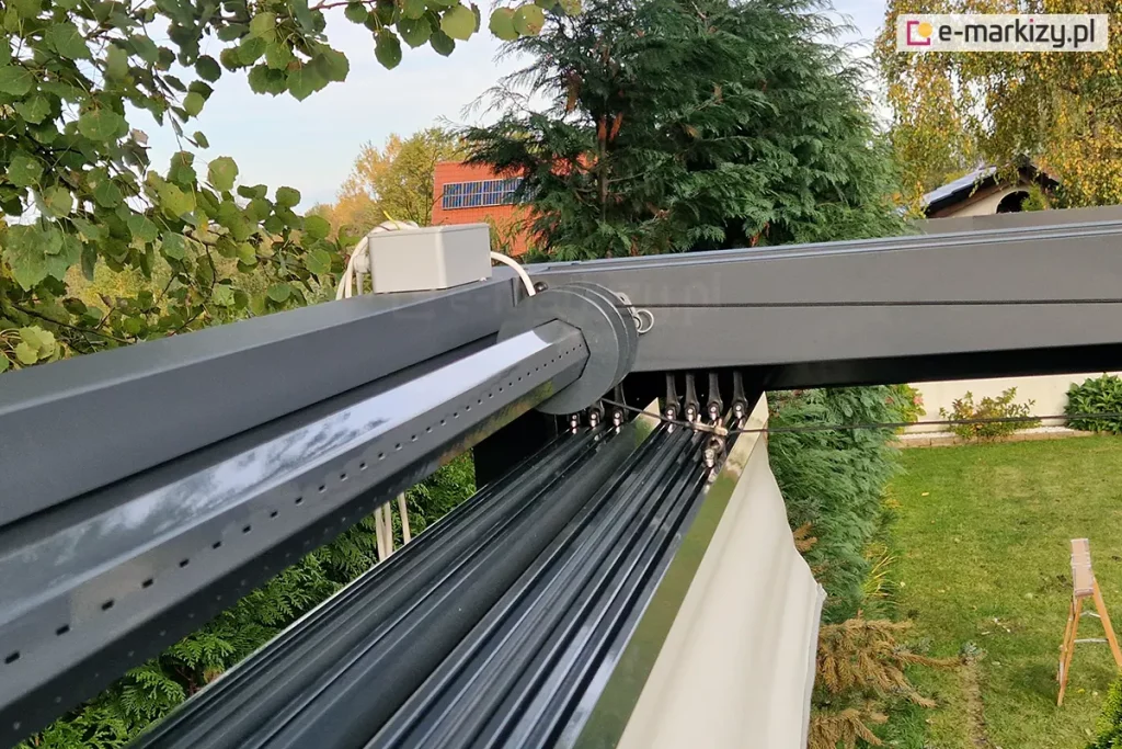 Narożnik konstrukcji pergoli Tako z widoczną rurą oktagonalną, w której ukryty pozostaje napęd oraz zainstalowanym na niej naciągiem linki. Do górnej części konstrukcji przymocowano skrzynkę w kolorze szarym z ukrytymi elementami sterującymi. Poniżej zwiniętego dachu widoczny zielony trawnik oraz liczne drzewa.