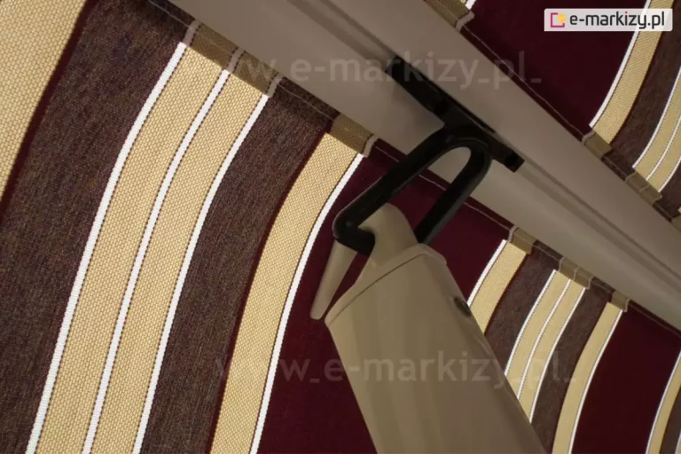 Belka przednia markizy balkonowej poręczowej z zamocowanym w jej rowku zaczepem oczkowym w kolorze czarnym. W zaczep wsunięty haczyk na końcu ramienia poręczowego, który zapewnia poszyciu odpowiednie naprężenie oraz wysięg. W tle widoczne poszycie w bordowe, beżowe, brązowe i białe pasy.