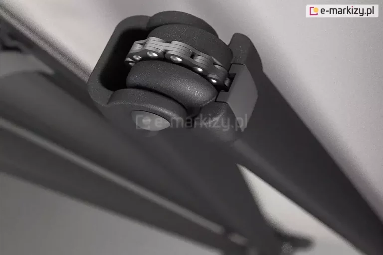 Detalu konstrukcyjny ramienia markizy tarasowej Aluprof Palladio, przedstawiający mechanizm przegubowy z wytrzymałym łańcuchem Flyera. Zdjęcie wykonane z bliskiej perspektywy ukazuje stalowy łańcuch wielopłytkowy osadzony na rolce przegubu, który odpowiada za przenoszenie sił naciągu sprężyn ukrytych wewnątrz aluminiowych profili ramienia. Ramię do markiz posiada przegub, którego konstrukcja wykonana jest z odlewanego aluminium malowanego proszkowo na kolor ciemnego grafitu o drobnej strukturze. Widoczne jest precyzyjne spasowanie elementów ruchomych oraz solidny sworzeń łączący, co zapewnia długowieczność mechanizmu i odporność na zmęczenie materiału przy częstym użytkowaniu markizy. Tło stanowi gładka powierzchnia rozłożonego poszycia akrylowego, co pozwala skupić uwagę na technicznym aspekcie napędu ramion przegubowych stosowanych w modelu Palladio.