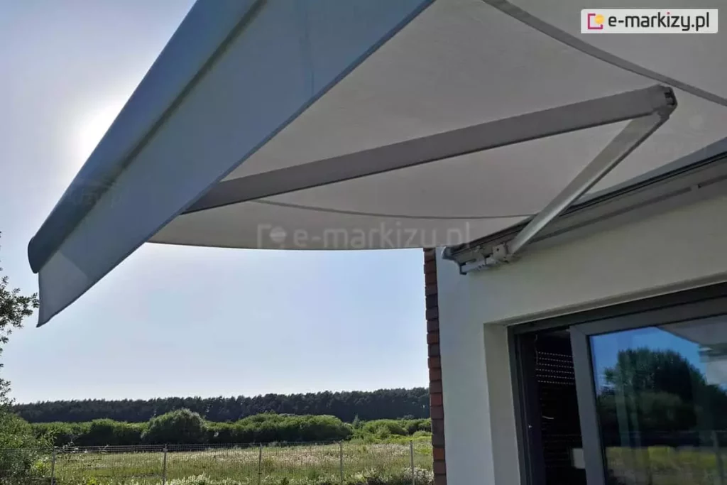 Zdjęcie wykonane w pełnym słońcu od dołu, przedstawiające rozłożoną markizę tarasową Palladio marki Aluprof zamontowaną nad przesuwnymi drzwiami tarasowymi nowoczesnego domu. Kadr skupia się na aluminiowym, przegubowym ramieniu naciągowym w kolorze jasnoszarym, które stabilizuje konstrukcję i napina gładkie, jasnoszare poszycie tkaniny. Przednia belka markizy została wyposażona w prostą, krótką falbanę o tym samym odcieniu, chroniącą przed promieniami słońca padającymi pod niskim kątem. Markiza przymocowana jest do białej elewacji z fragmentem narożnika wykończonego ciemną cegłą klinkierową. W tle, pod zadaszeniem, rozpościera się widok na zielony ogród, ogrodzenie z siatki, łąki oraz gęstą linię lasu na horyzoncie pod bezchmurnym, błękitnym niebem. Promienie słoneczne przebijają się zza lewej krawędzi poszycia, tworząc naturalną poświatę i podkreślając ochronną funkcję zamontowanej osłony.