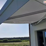 Zdjęcie wykonane w pełnym słońcu od dołu, przedstawiające rozłożoną markizę tarasową Palladio marki Aluprof zamontowaną nad przesuwnymi drzwiami tarasowymi nowoczesnego domu. Kadr skupia się na aluminiowym, przegubowym ramieniu naciągowym w kolorze jasnoszarym, które stabilizuje konstrukcję i napina gładkie, jasnoszare poszycie tkaniny. Przednia belka markizy została wyposażona w prostą, krótką falbanę o tym samym odcieniu, chroniącą przed promieniami słońca padającymi pod niskim kątem. Markiza przymocowana jest do białej elewacji z fragmentem narożnika wykończonego ciemną cegłą klinkierową. W tle, pod zadaszeniem, rozpościera się widok na zielony ogród, ogrodzenie z siatki, łąki oraz gęstą linię lasu na horyzoncie pod bezchmurnym, błękitnym niebem. Promienie słoneczne przebijają się zza lewej krawędzi poszycia, tworząc naturalną poświatę i podkreślając ochronną funkcję zamontowanej osłony.