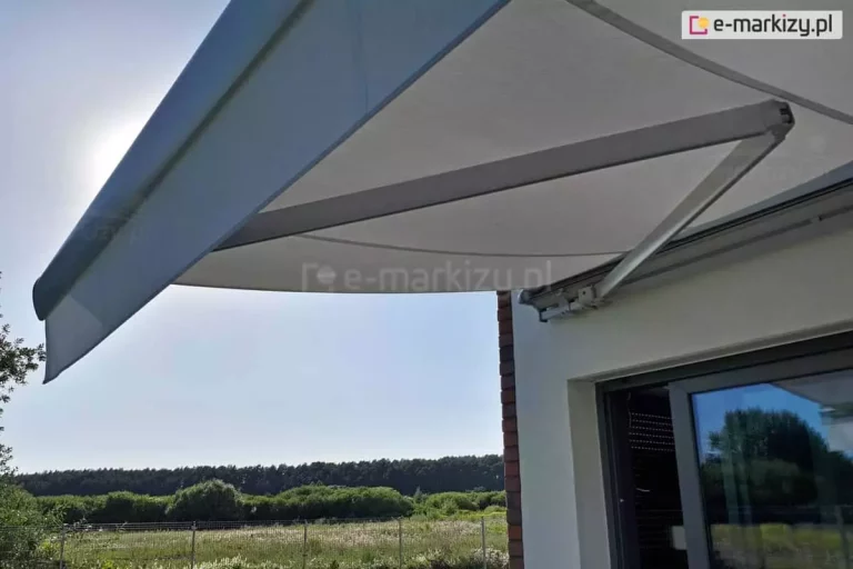 Zdjęcie wykonane w pełnym słońcu od dołu, przedstawiające rozłożoną markizę tarasową Palladio marki Aluprof zamontowaną nad przesuwnymi drzwiami tarasowymi nowoczesnego domu. Kadr skupia się na aluminiowym, przegubowym ramieniu naciągowym w kolorze jasnoszarym, które stabilizuje konstrukcję i napina gładkie, jasnoszare poszycie tkaniny. Przednia belka markizy została wyposażona w prostą, krótką falbanę o tym samym odcieniu, chroniącą przed promieniami słońca padającymi pod niskim kątem. Markiza przymocowana jest do białej elewacji z fragmentem narożnika wykończonego ciemną cegłą klinkierową. W tle, pod zadaszeniem, rozpościera się widok na zielony ogród, ogrodzenie z siatki, łąki oraz gęstą linię lasu na horyzoncie pod bezchmurnym, błękitnym niebem. Promienie słoneczne przebijają się zza lewej krawędzi poszycia, tworząc naturalną poświatę i podkreślając ochronną funkcję zamontowanej osłony.